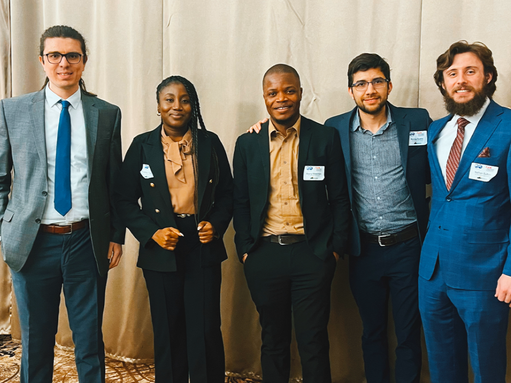 Five graduate students in business attire in front of golden curtain backdrop.