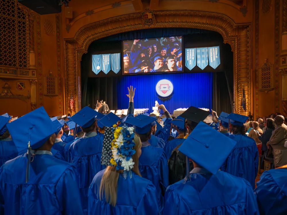 A view of the stage decorated for commencement over a sea of students wearing blue graduation mortarboard caps