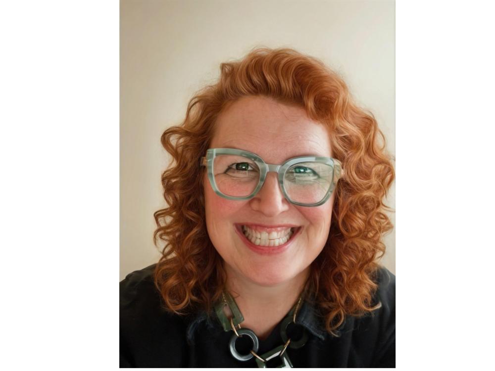 headshot of smiling person with curly red hair, green glasses, chunky necklace