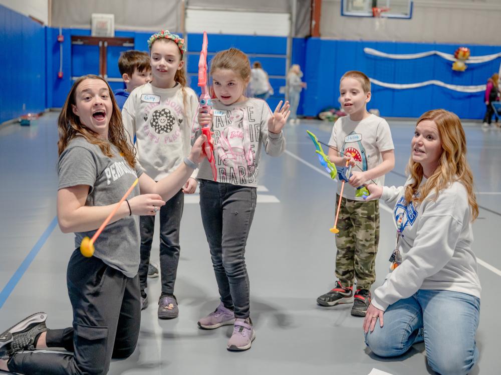 A couple of students help some children with a bow and arrow in a gymnasium
