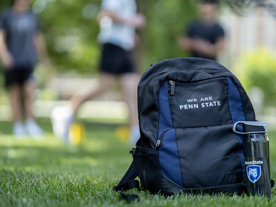 A backpack with the words "We Are Penn State" next to a water bottle with the Penn State logo sitting on a grassy field.