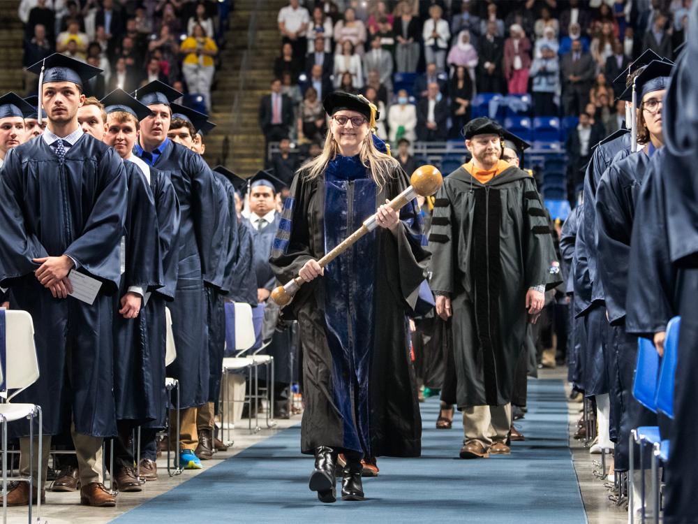 Person walking down aisle at commencement, surrounded by others watching on.