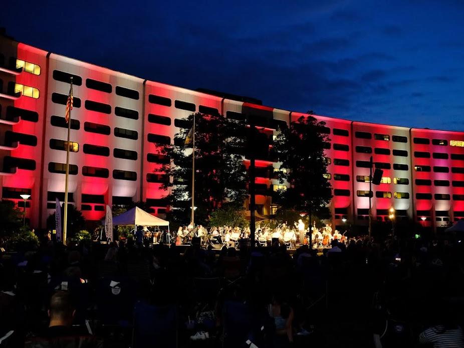 Penn State Health Milton S. Hershey Medical Center is lit up in red and white lights as an orchestra plays on a lit stage in front of the building's signature Crescent. A crowd of a couple thousand people looks on.