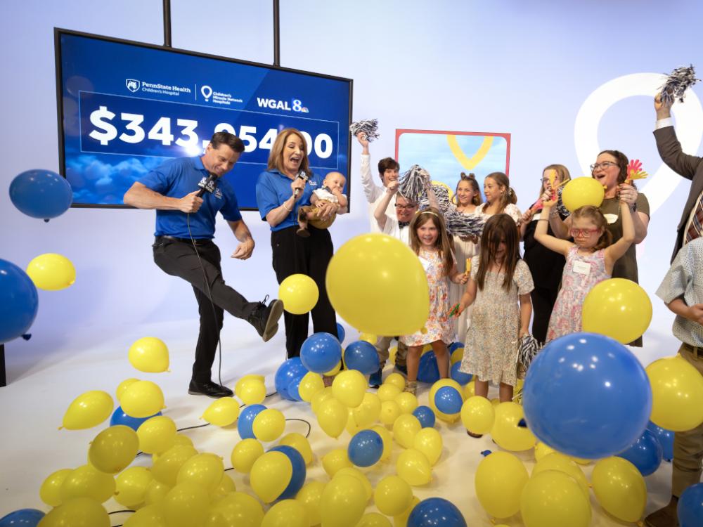 A group of excited children and adults celebrate on stage as blue and yellow balloons fall around them. Two WGAL 8 news anchors, one holding a baby, cheer in front of a large screen displaying a fundraising total of $343,054.00.
