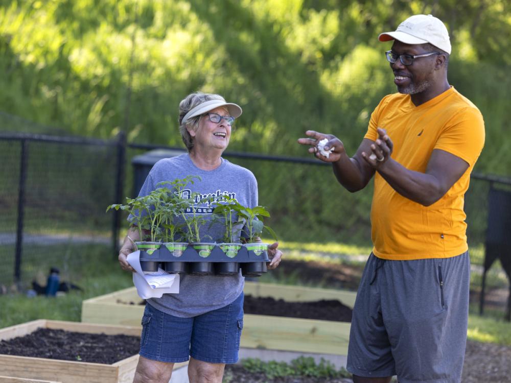 A man and a woman stand side-by-side talking in a community garden. The woman is holding a container with several potted plants. Both individuals are smiling.