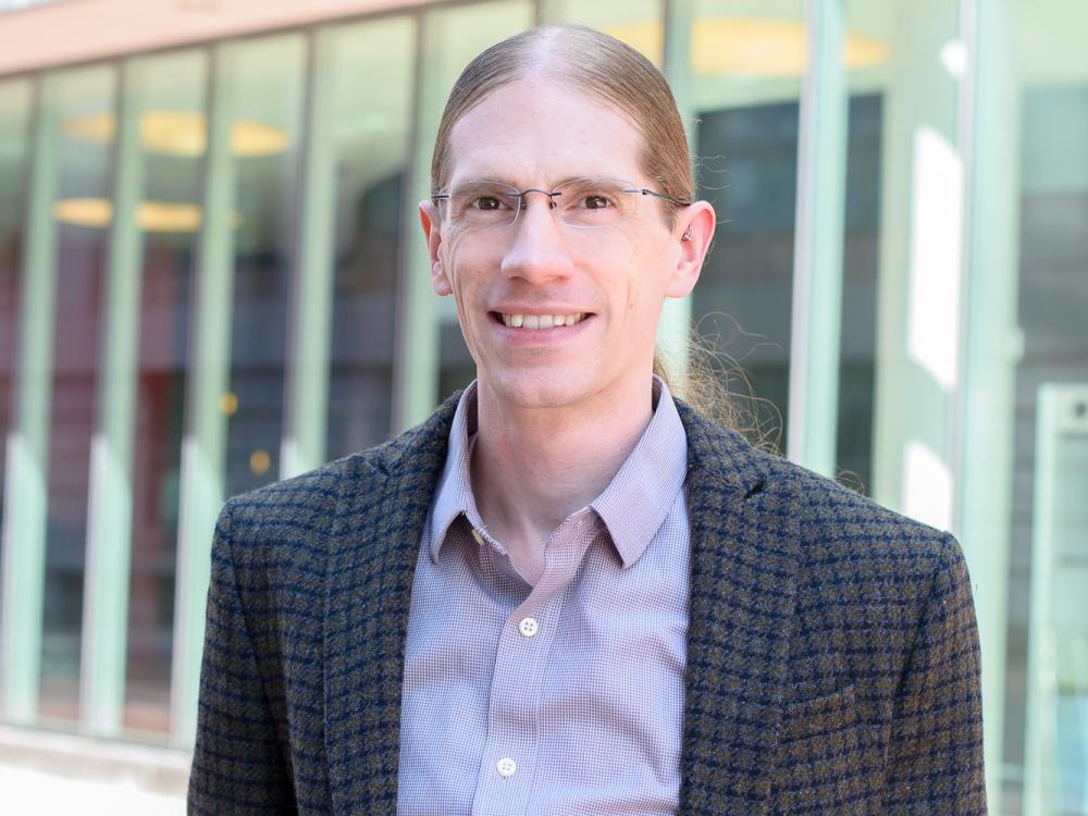 A person with long hair pulled back in glasses and a purple shirt with a plaid blazer smiles at the camera.
