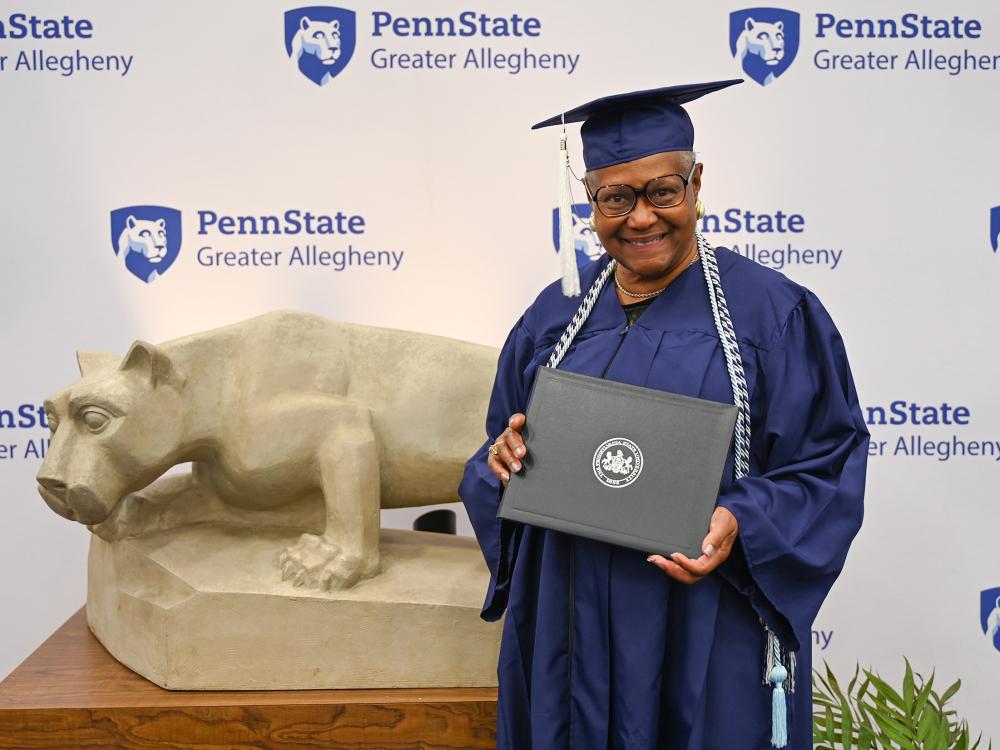 Diane Banks, wearing a graduation cap and gown, smiles while holding her diploma in front of the Penn State Greater Allegheny backdrop and Nittany Lion statue.