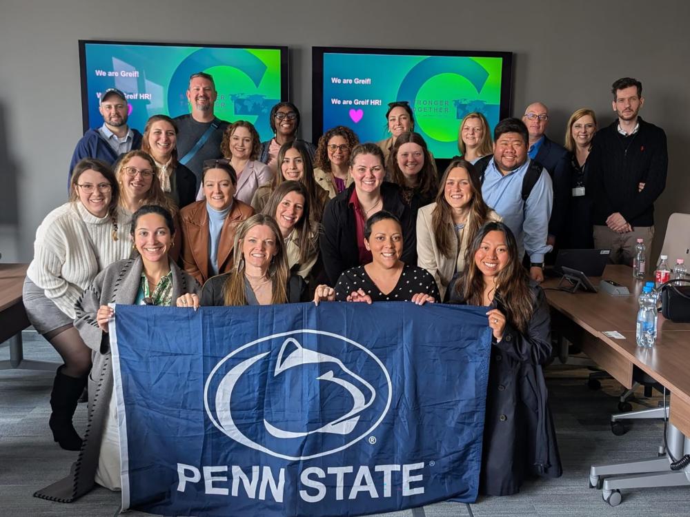 A group of people pose with a blue Penn State flag in a conference room.