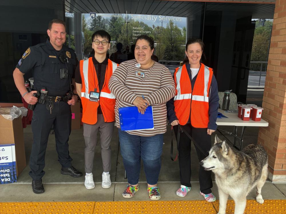 A police officer, three colleagues and a dog stand next to tables and collection boxes in front of a medical practice.
