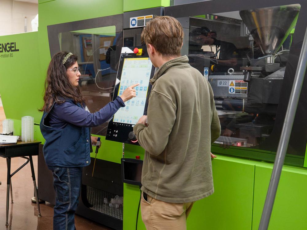 A person operates a bright green injection molding machine while another person looks on.