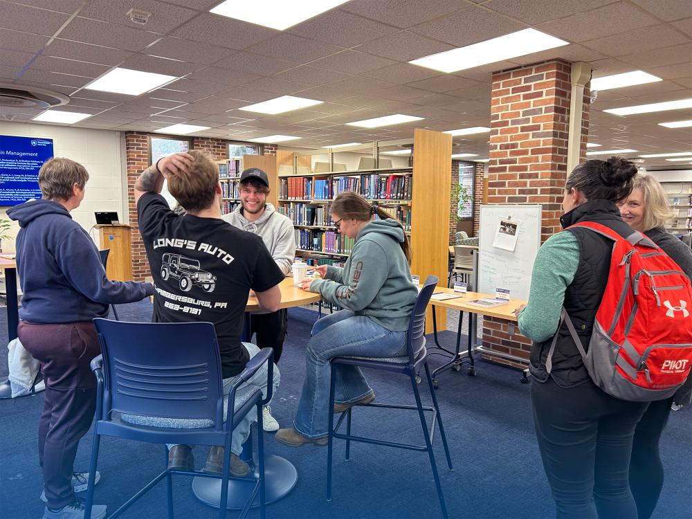 Four people sitting around a table talking and laughing. Two additional people standing to the right talking and laughing.