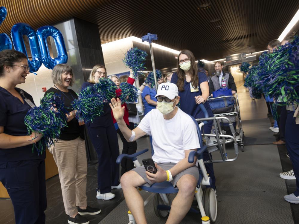 A man wearing a white t-shirt, white baseball cap, and face mask sits in a wheelchair and waves while holding a phone in his lap. He is surrounded by smiling hospital staff wearing scrubs and holding blue and green pom-poms.