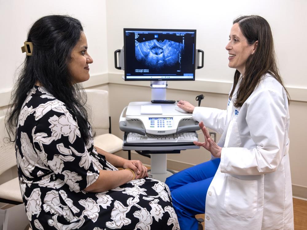 Sushrutha Sridhar, left, talks to Stephanie Estes of Penn State Health Obstetrics and Gynecology in a doctor’s office. Sridhar is wearing a flowered dress, and Estes is wearing a white lab coat. An ultrasound image of a uterus is shown on a screen between them.