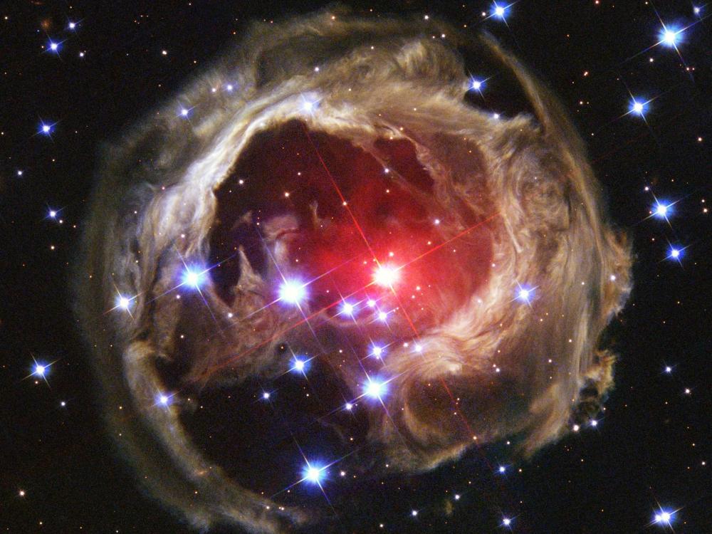 Space image — at the center, a bright red star is surrounded by a circular cloud of gas and dust with intricate patterns. Numerous blue and white stars are scattered across the black background of space.