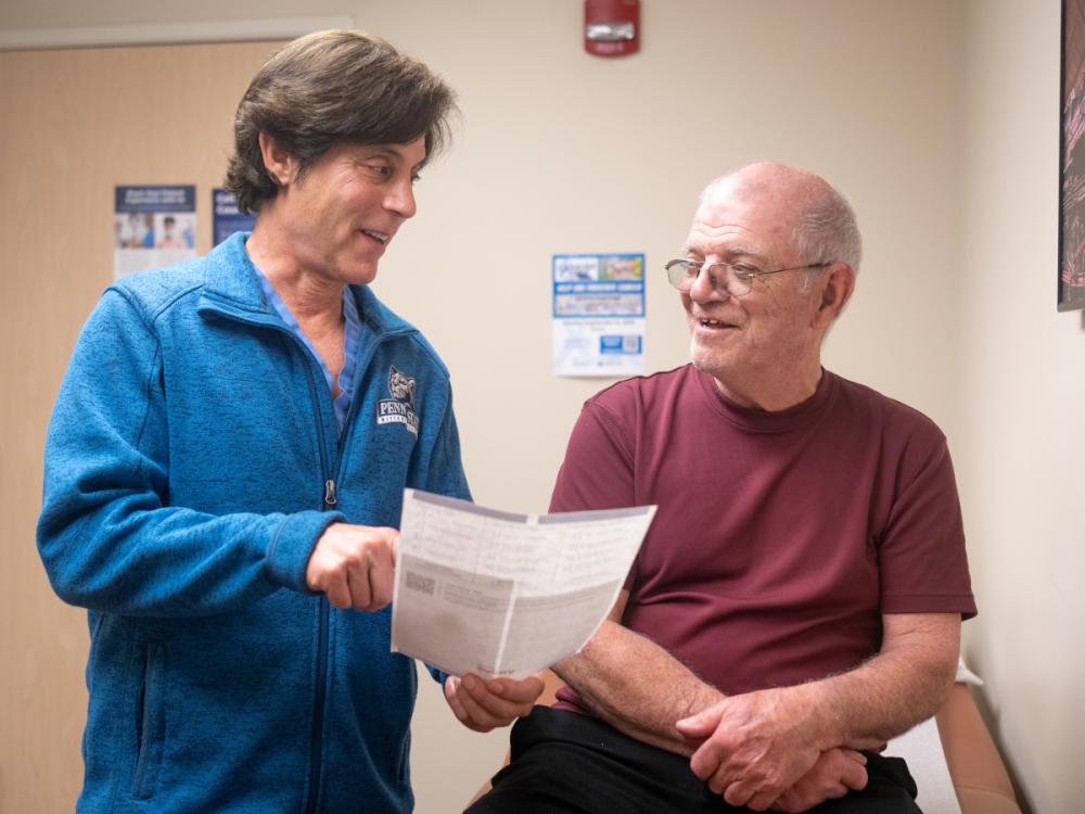 A doctor smiles at a male patient as he points to a paper he is holding. The doctor is wearing a sweat jacket with a Penn State logo on it. The male is wearing a T-shirt and glasses.