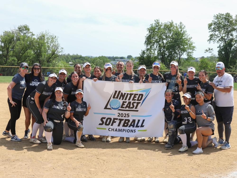Penn State Brandywine's softball team holds the championship banner on the softball field.