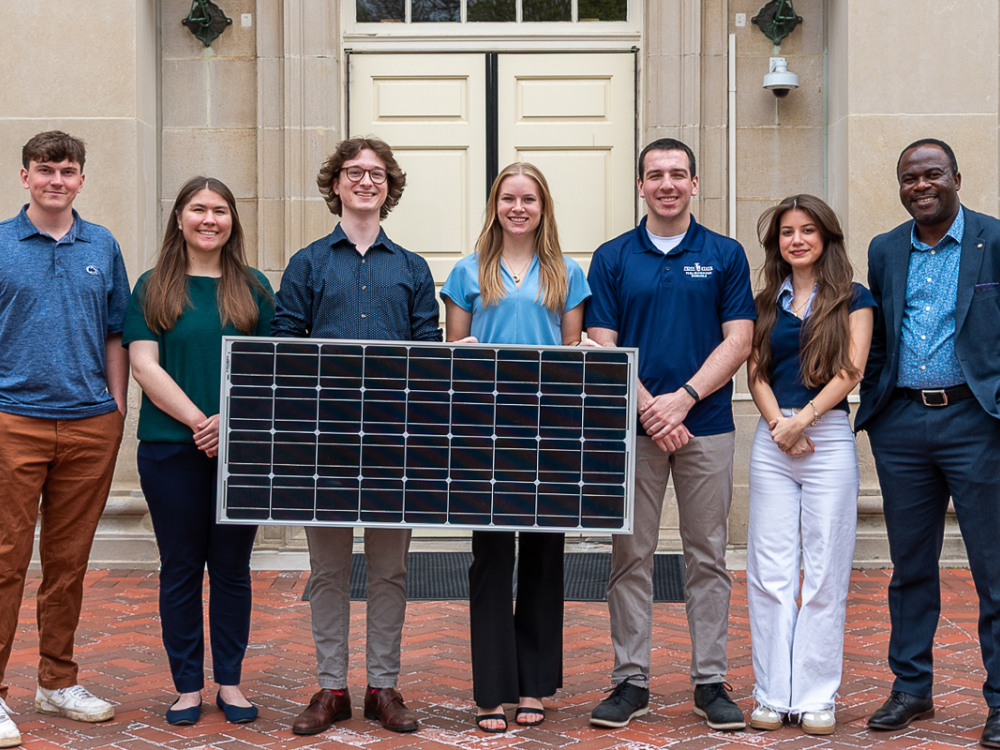 Student group stands in front of Hosler Building holding a solar panel. 