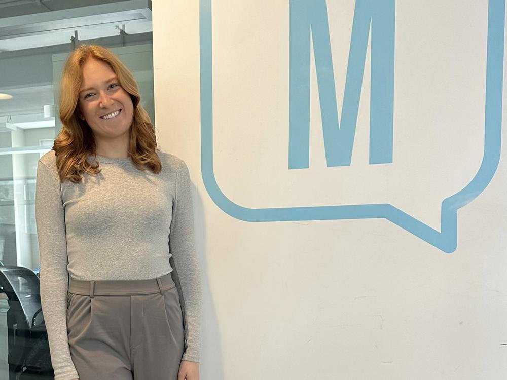 Smiling long-haired girl standing by a wall with an "M' logo