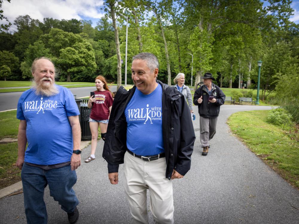 Two men wearing Walk With a Doc T-shirts smile as they walk at City Park in Reading. Behind them, three other people walk along the path.