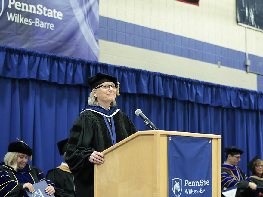 A woman in academic regalia speaks at a podium bearing the Penn State Wilkes-Barre logo.