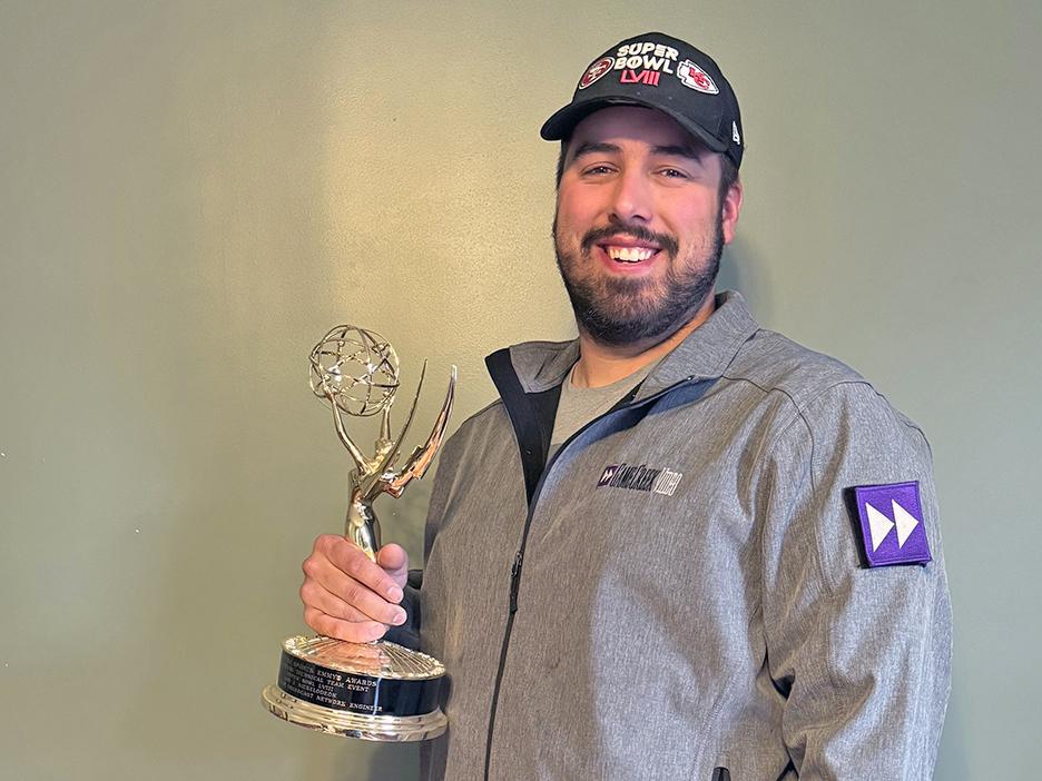 A man in a black baseball cap and gray jacket holding an Emmy statuette.