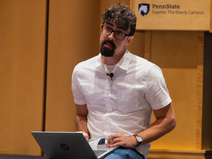 Allain Daigle sits with a laptop in his lap, speaking during a faculty development session at Penn State Fayette. A Penn State logo and campus mark are visible in the background.