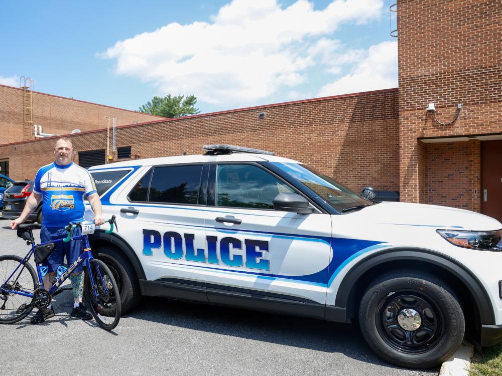 Bill Carter in a Delco 476ers bike jersey standing behind his bike, in front of Penn State University Police car