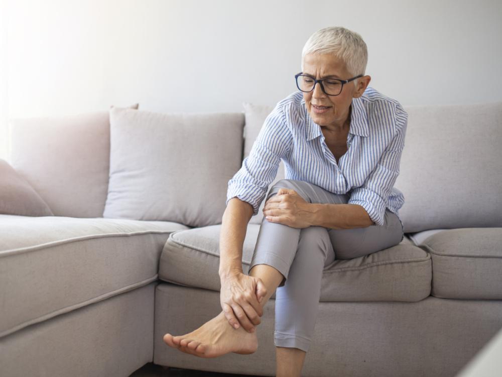 An older woman suffering from leg pain holds her ankle at home.