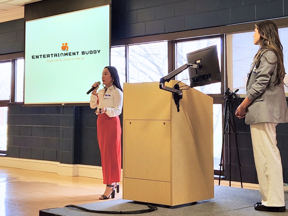 Two individuals stand presenting in front of a screen that reads Entertainment Buddy
