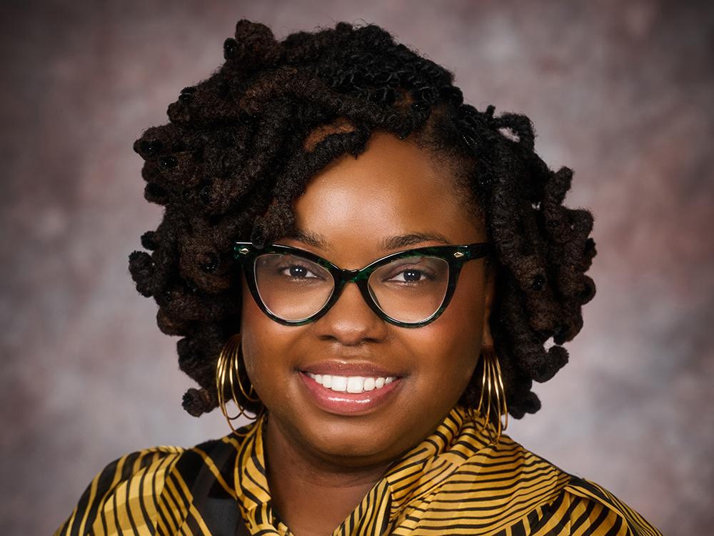 Portrait of Mariah Harmon in front of gray background. She is wearing glasses and a black-and-yellow shirt.