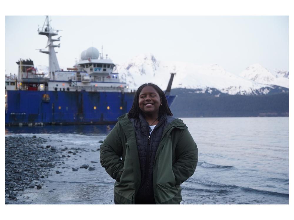 young adult stands in front of large ship against alaska skyline