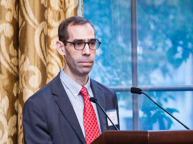 A man with glasses wearing a blue suit and red tie stands behind a podium.