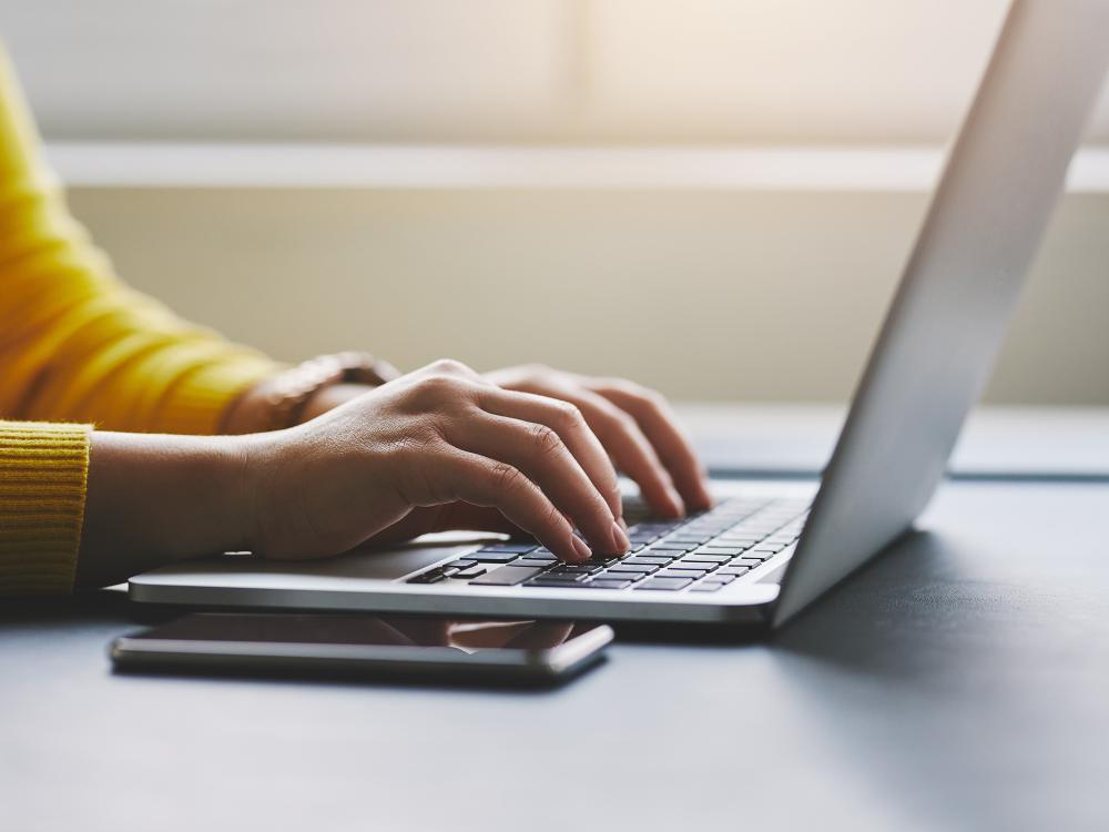 A person wearing a yellow shirt types on a laptop. Only the person's hands and arms are visible.