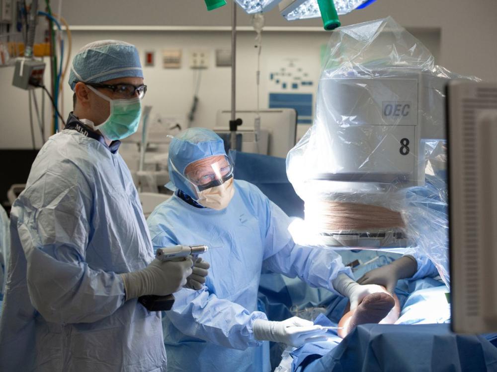 Medical professionals in full surgical attire perform a procedure in an operating room. One holds a surgical drill while another works on a patient's leg.