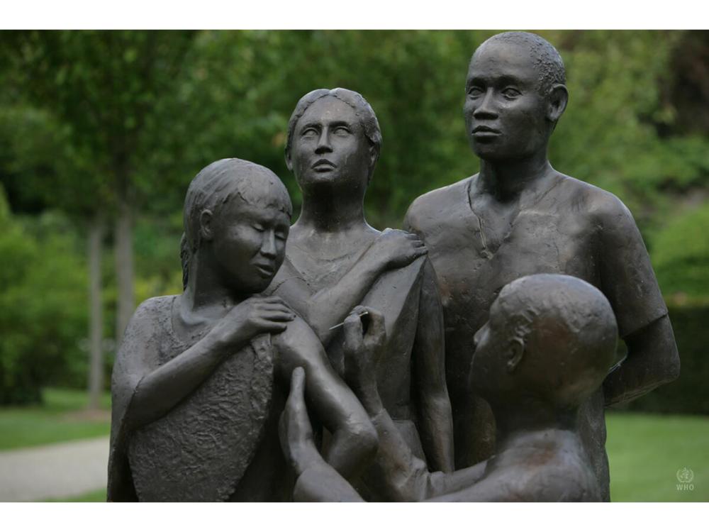 Bronze statue of health worker giving vaccine to one person as two others look on.