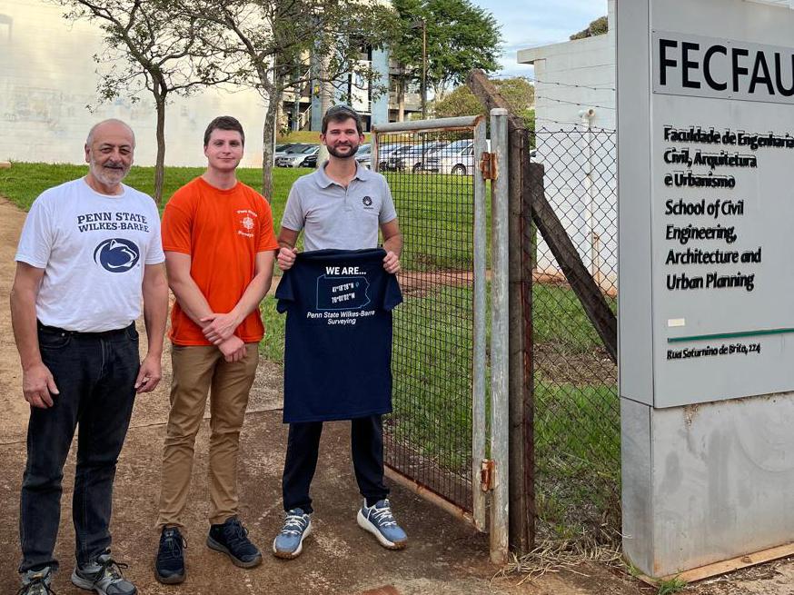 Three people, one holding a Penn State Wilkes-Barre shirt, stand outside a building in Brazil.