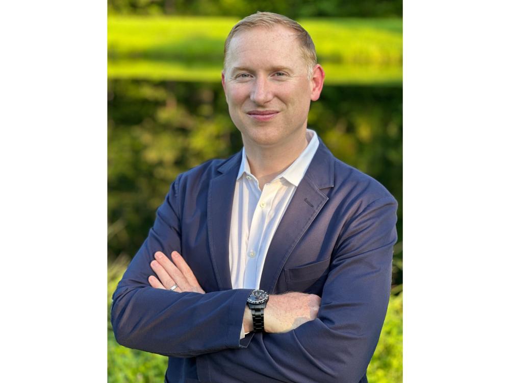 fair-haired person in white shirt and blue sportcoat stands with arms folded