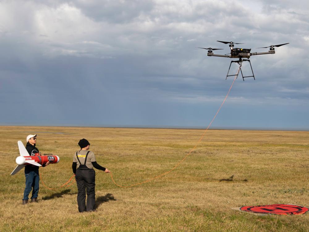 People standing outside, watching drone fly up into the Alaskan air. Green grass all around with no mountains of any kind in the background.