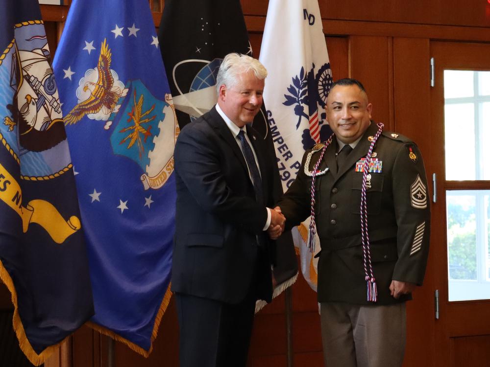 a man in a suit shakes hand with an Army sergeant major wearing a brown uniform. they're standing on a stage with military flags in the background.
