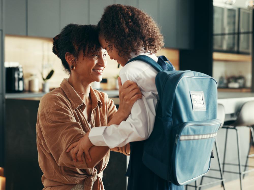 Mom and girl touch foreheads as girl gets ready for school. Girl is wearing a school uniform and backpack.