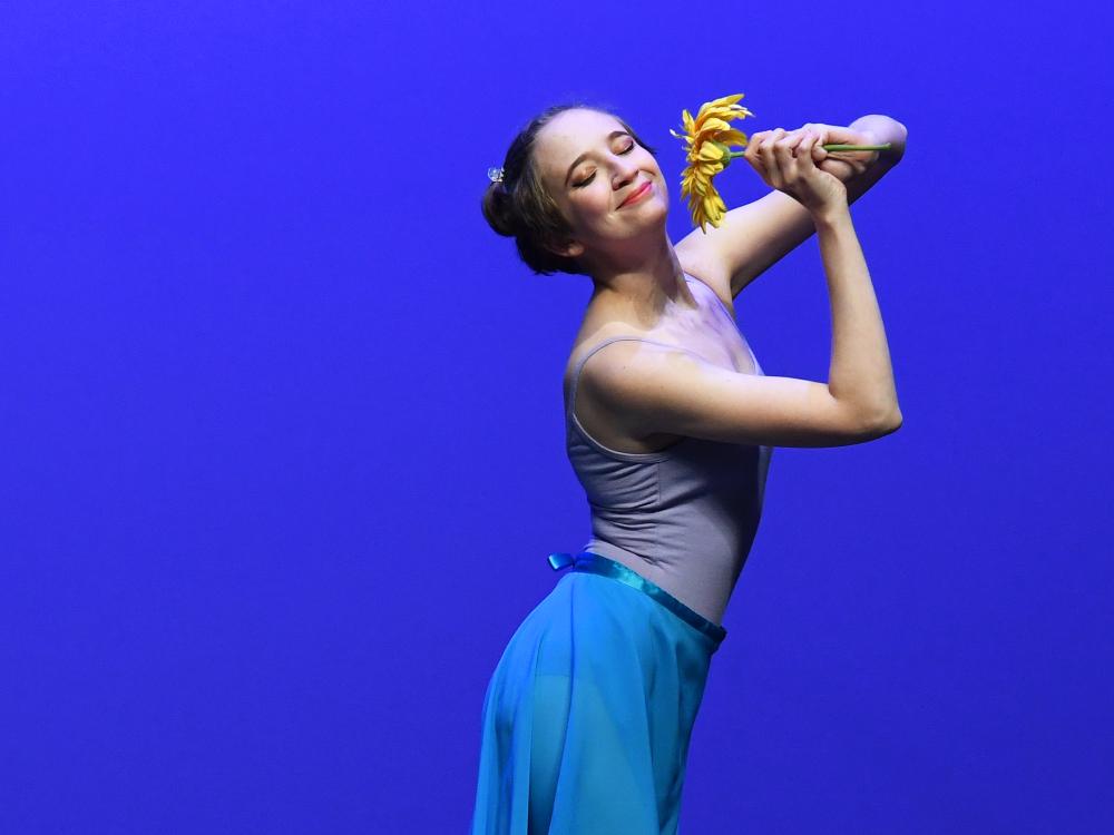 A ballet dancer in a lavender leotard and flowing blue skirt poses gracefully on stage, holding a yellow flower near her face with a joyful expression against a solid blue background.
