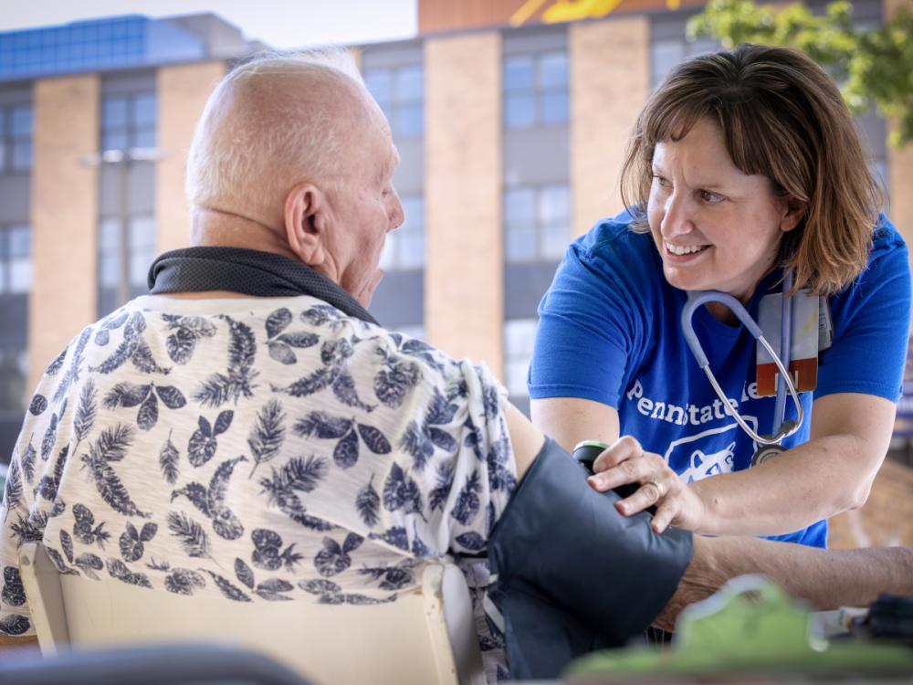 A community health nurse at Penn State Health smiles at a man as she checks his blood pressure. She is wearing a T-shirt and has a stethoscope around her neck. He is wearing a shirt with a leaf print on it.