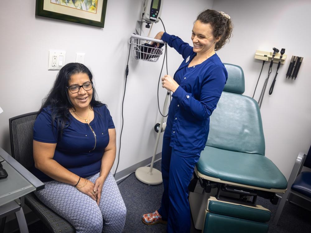 A woman patient smiles as she sits in a chair in a doctor’s exam room, while a nurse in scrubs puts a blood pressure cuff in a basket on the wall.