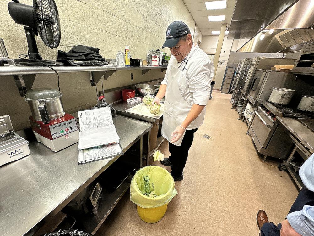 A man in a white chefs jacket throws cauliflower stems into a yellow composting bin
