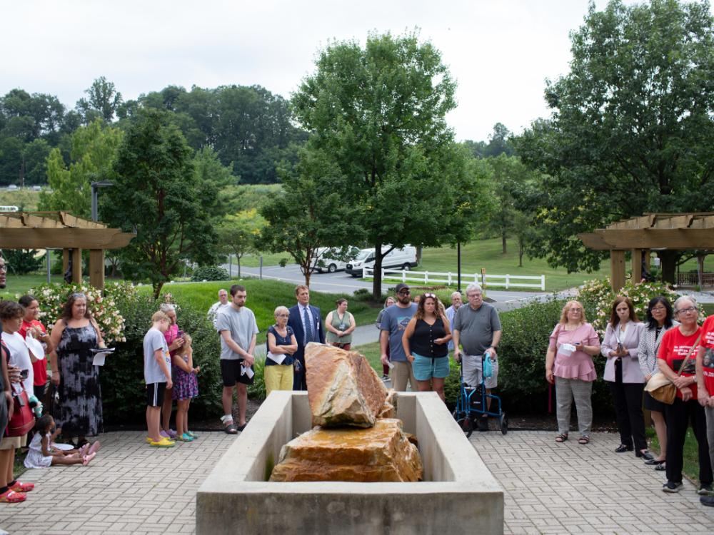 Approximately 20 people stand on a brick patio, surrounding a stone memorial, at a ceremony.