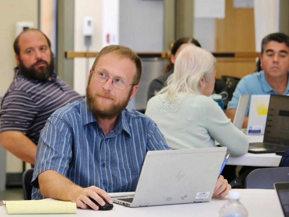 An individual sits at table with other table groupings all working on laptops