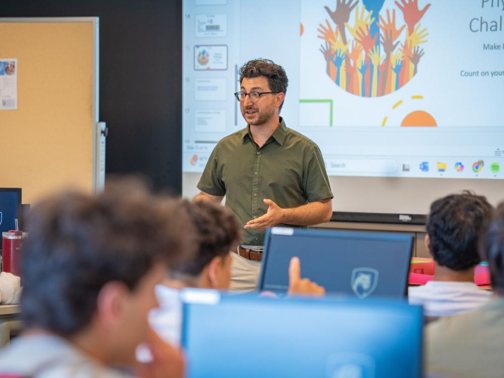 college professor with dark hair and glasses speaks to class