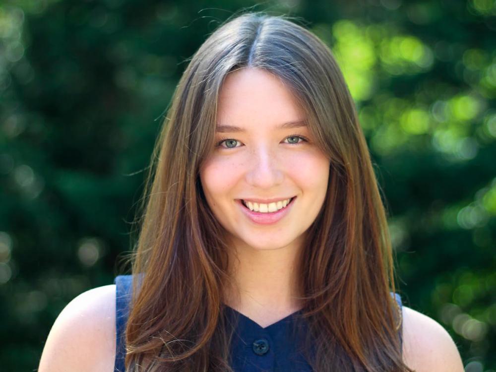 Kathryn Kearney who has long dark brown hear and is wearing a blue button-down sleeveless shirt, stands outside.