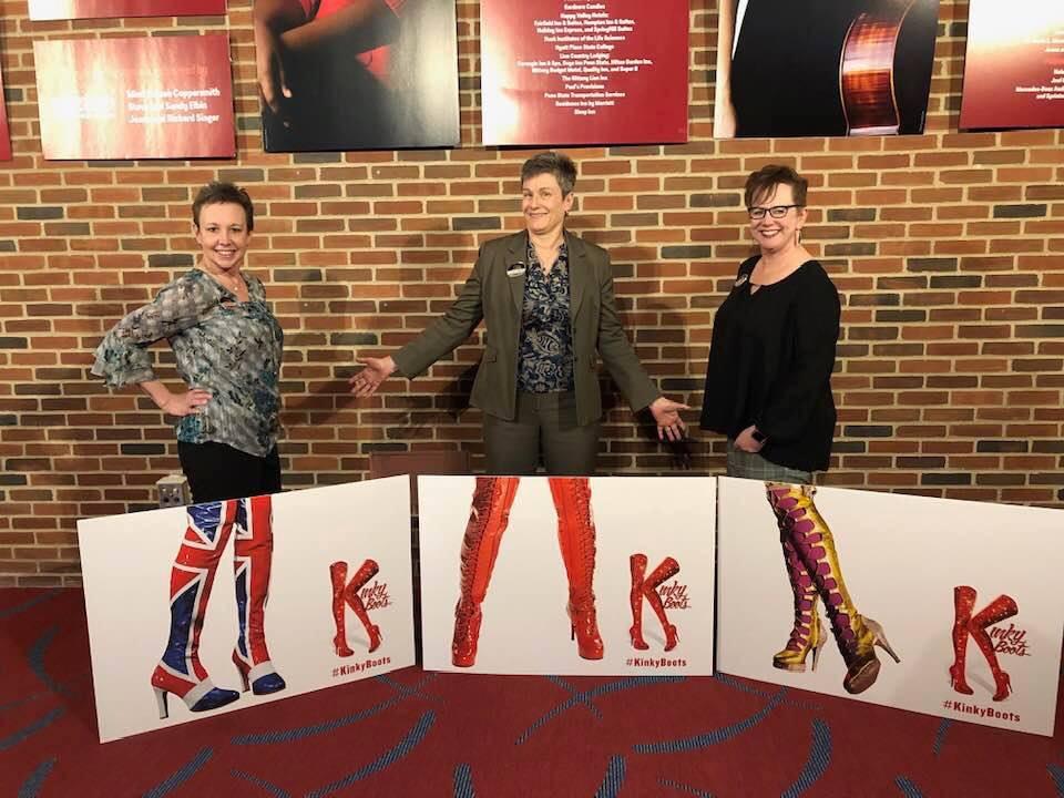 Three women stand in front of cardboard cutouts resembling stiletto boots.