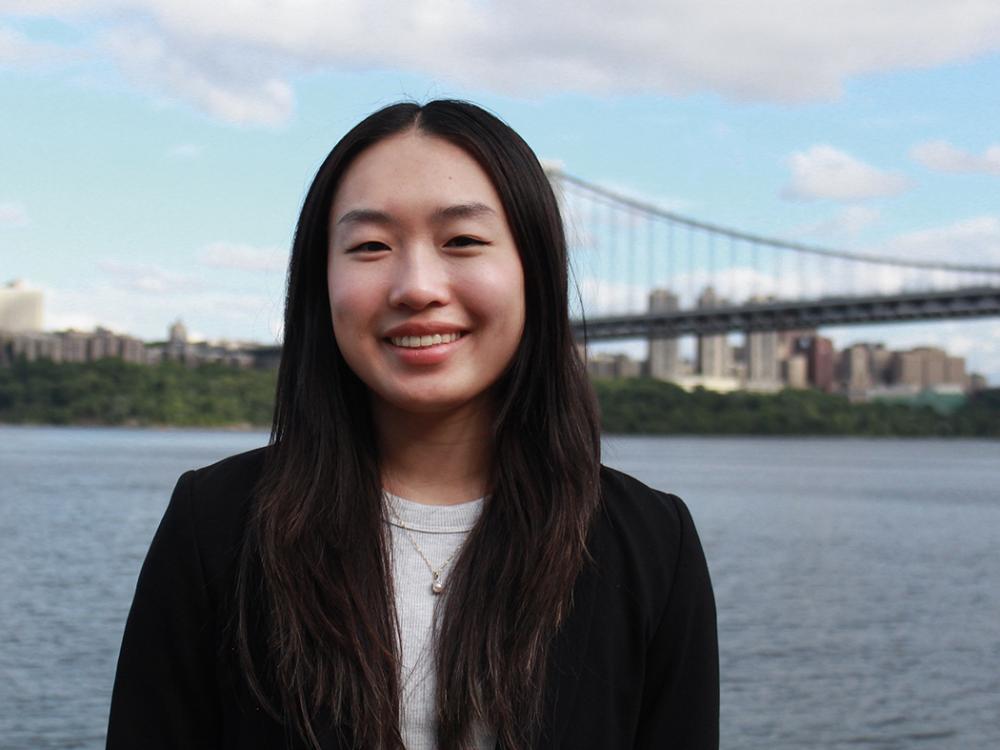Photo of dark haired student with black blazer and white shirt with a river and bridge in background.